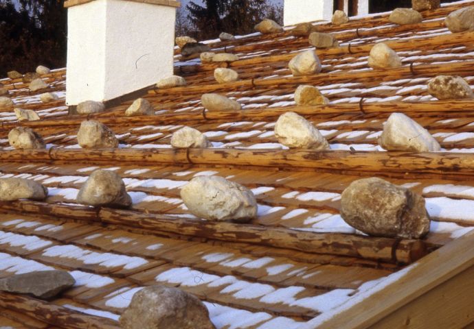 Erster Schnee auf dem frisch gedeckten Knotzingerhaus, 1980. Blick auf das Legeschindeldach mit zwei Kaminen. Die Schindeln sind mit Steinen gesichert, die auf querliegenden Holzstangen liegen.