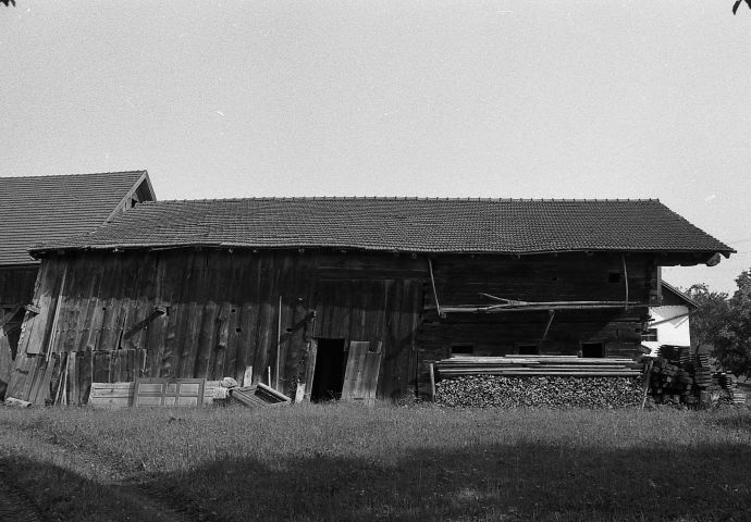 Das Haus an seinem ursprünglichen Standort in Helming. Ansicht auf eine Längsseite mit Holzstoß rechts. Ein Trockengestell hängt über dem Holzstoß..
