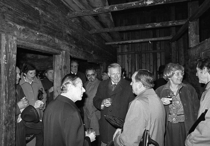 Kurt Conrad mit Gästen bei der Firstfeier des Zischkhäusls, 1979. Kurt Conrad ist umringt von den Gästen, er spricht mit zwei Herren. Die Szene findet innen im Haus statt.