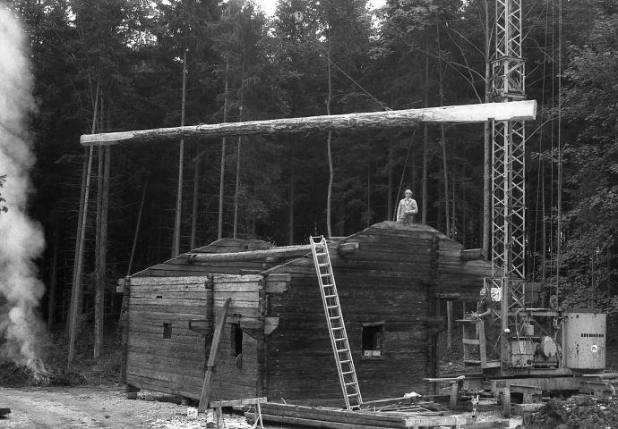 Beim Aufbau im Freilichtmuseum, 1984. Seitenansicht auf die Tenne. Sie steht an ihrem Platz, noch kein Dach. An die Hauswand lehnt eine Leiter. Links vom Haus brennt ein Feuer Der Rauch steigt gerade hoch. Rechts der untere Teil eines Krans.