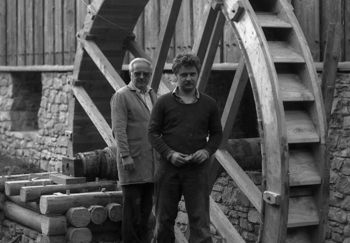 Beim Aufbau im Freilichtmuseum, 1984. Zwei Mitarbeiter stehen vor dem Wasserrad. Es ist bereits aufgebaut. Bis ca. zur Hälfte der Höhe des Wasserrades ist das Steinfundament sichtbar.