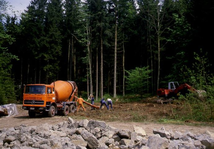Bei der Abtragung in Gumping, 1984. Links im Bild ein roter Betonmischer, in der Mitte wird das Fundament gegossen. Rechts im Bild ein Traktor. Wald im Hintergrund.