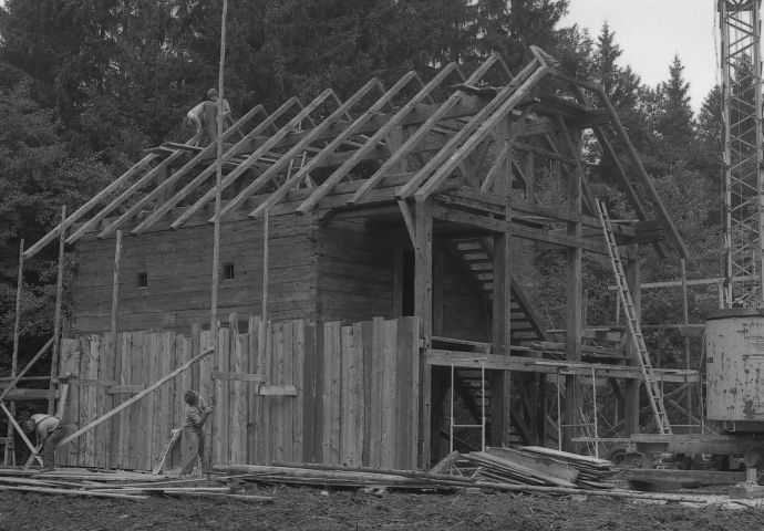 Beim Aufbau im Freilichtmuseum, 1984. Das Holzgerüst inklusive Dachstock steht. Man sieht den Innenkasten der Meindlhütte. Arbeiter verkleiden die Aussenwand mit Holzbrettern. Die Dachdeecker sind auch an der Arbeit.