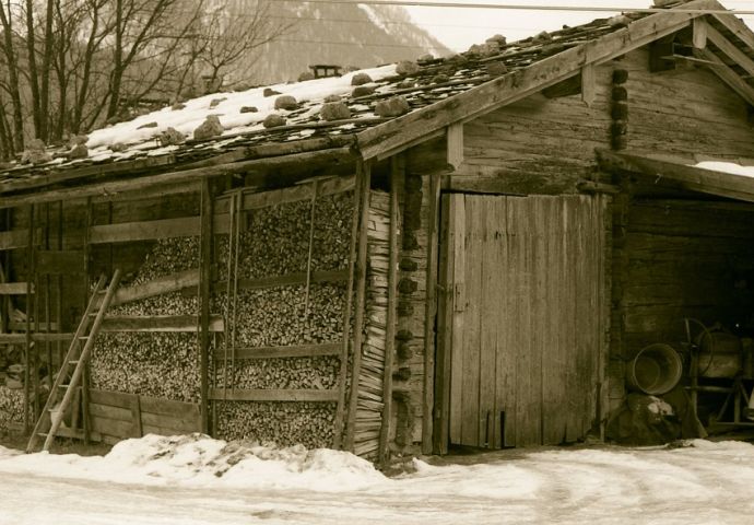 Die Getreidetenne an ihrem ursprünglichen Standort in Gumping. Seitliche Ansicht im Winter. An der linken Längsseite Holzstöße bis unters Dach.  Das Tor auf der Fronsteite ist zu. Schnee liegt auf dem Dach. Es ist eine Legeschindeldach.