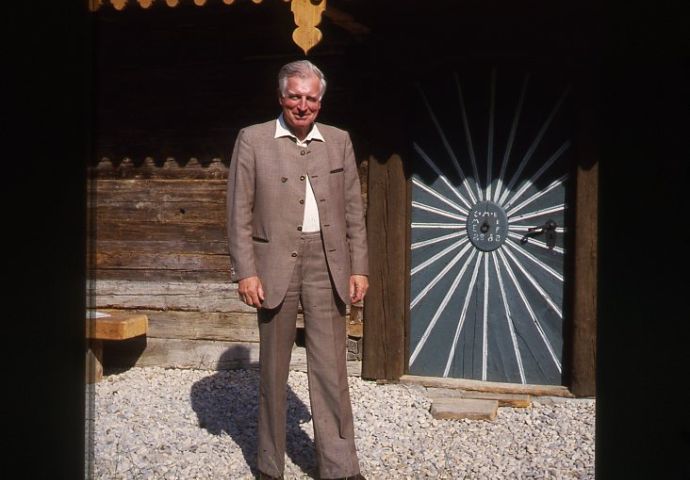 Kurt Conrad vor der Haustüre. Kurt Conrad steht links von der markanten Sonnentüre. Auf dem grünen Hintergrund zeichnet sich in weiß die Sonne mit den Strahlen ab. ist das Dekor der Balkonverzierung sichtbar.,