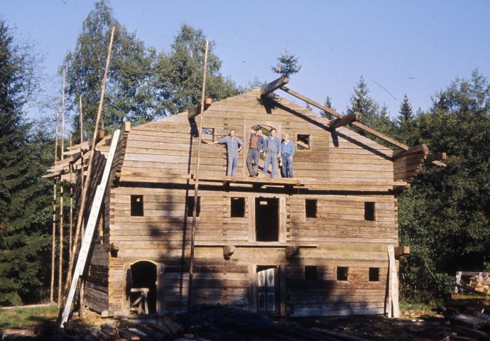 Beim Aufbau im Salzburger Freilichtmuseum, 1984. Die Firstfeier hat bereits stattgefunden, das kleine Bäumchen ist noch sichtbar. Frontalansicht. Vier Arbeiter stehen auf dem Balkonbalken im zweiten Stockwerk.