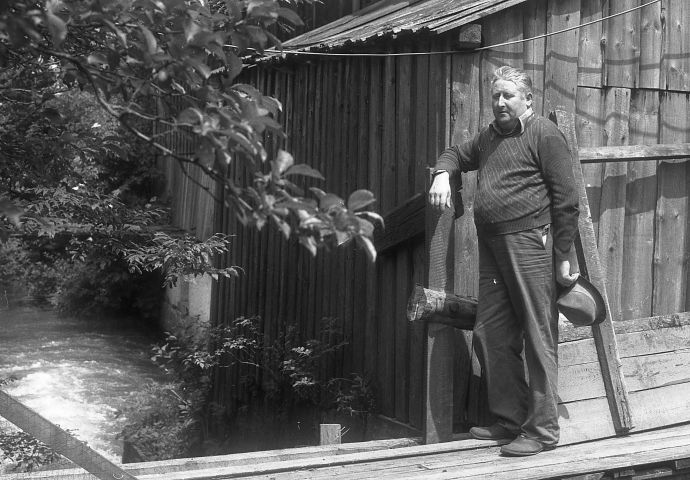 Der Vorbesitzer der Mittermühlsäge, 1980. Er steht auf einer schmalen Holzbrücke und lehnt sich an einen Holzpfahl vor einem Schopf.