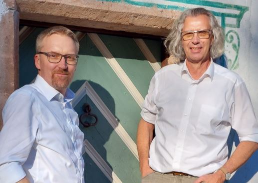 links steht Peter Fritz, zukünftiger Direktor. Rechts steht Michael Weese, aktueller Direktor des Salzburger Freilichtmuseums. Sie stehen in einer Türe und lehnen entspannt an den jeweiligen Türpfosten.
